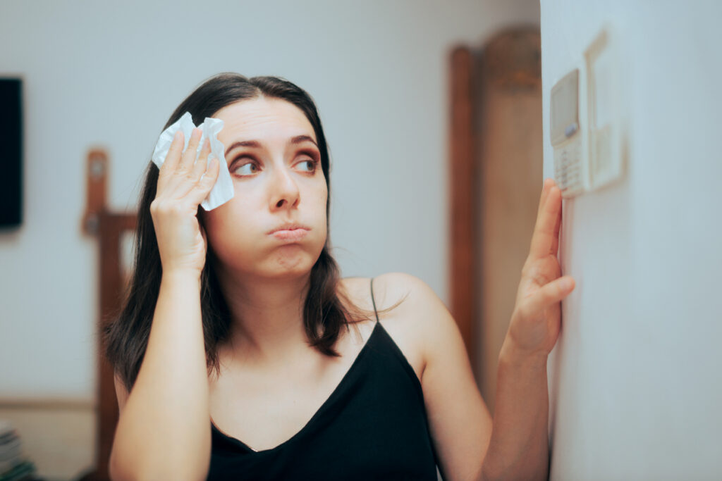 Woman wiping sweat off her forehead as she checks a thermostat on a white wall.