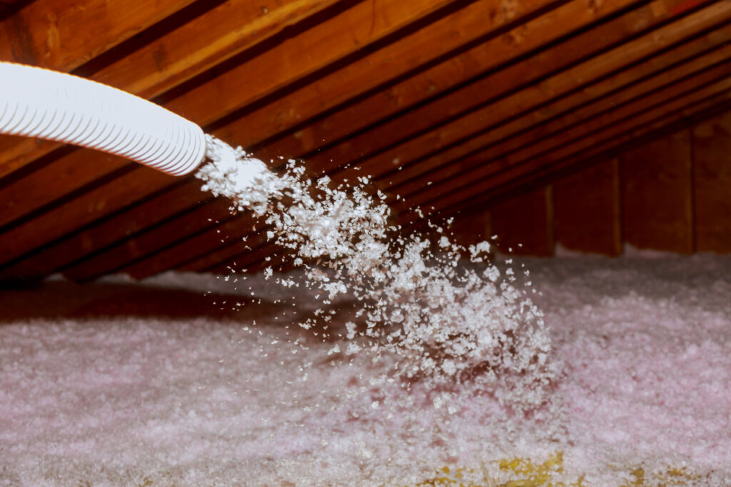 Plural component gun spraying white blown fiberglass insulation into an attic.