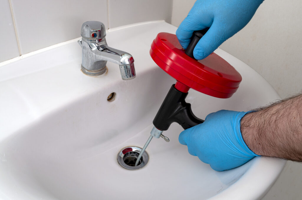 Close view of a plumber's hands in blue latex gloves using a drain snake to clear a bathroom sink drain.