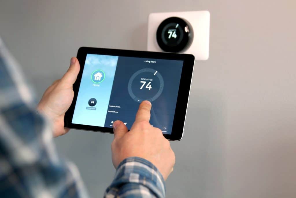 Person adjusting the temperature using a tablet with smart home app in modern living room. Smart thermostat on light gray wall in background.