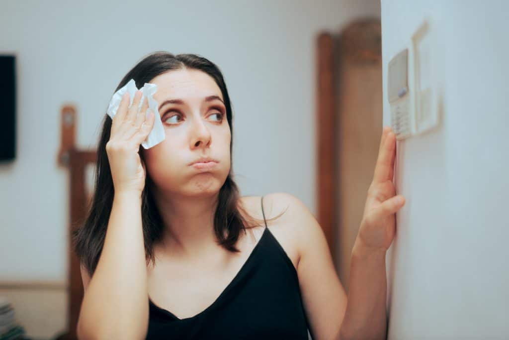 Woman wiping sweat from her forehead as she checks the thermostat on her wall.